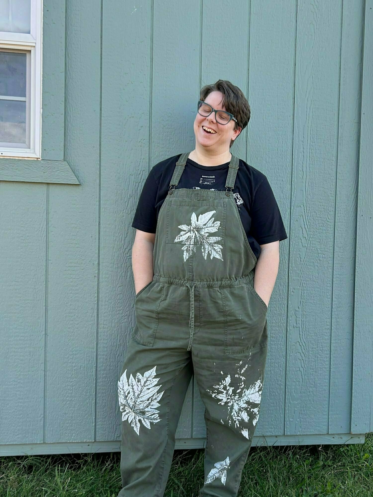 I'm a nonbinary human with pale skin, green glasses, and short brownish hair caught on camera laughing by my then four year old. I am wearing hand printed overalls with giant white leaves and a black shirt. My hands are in my pockets and my gray green art studio is behind me.