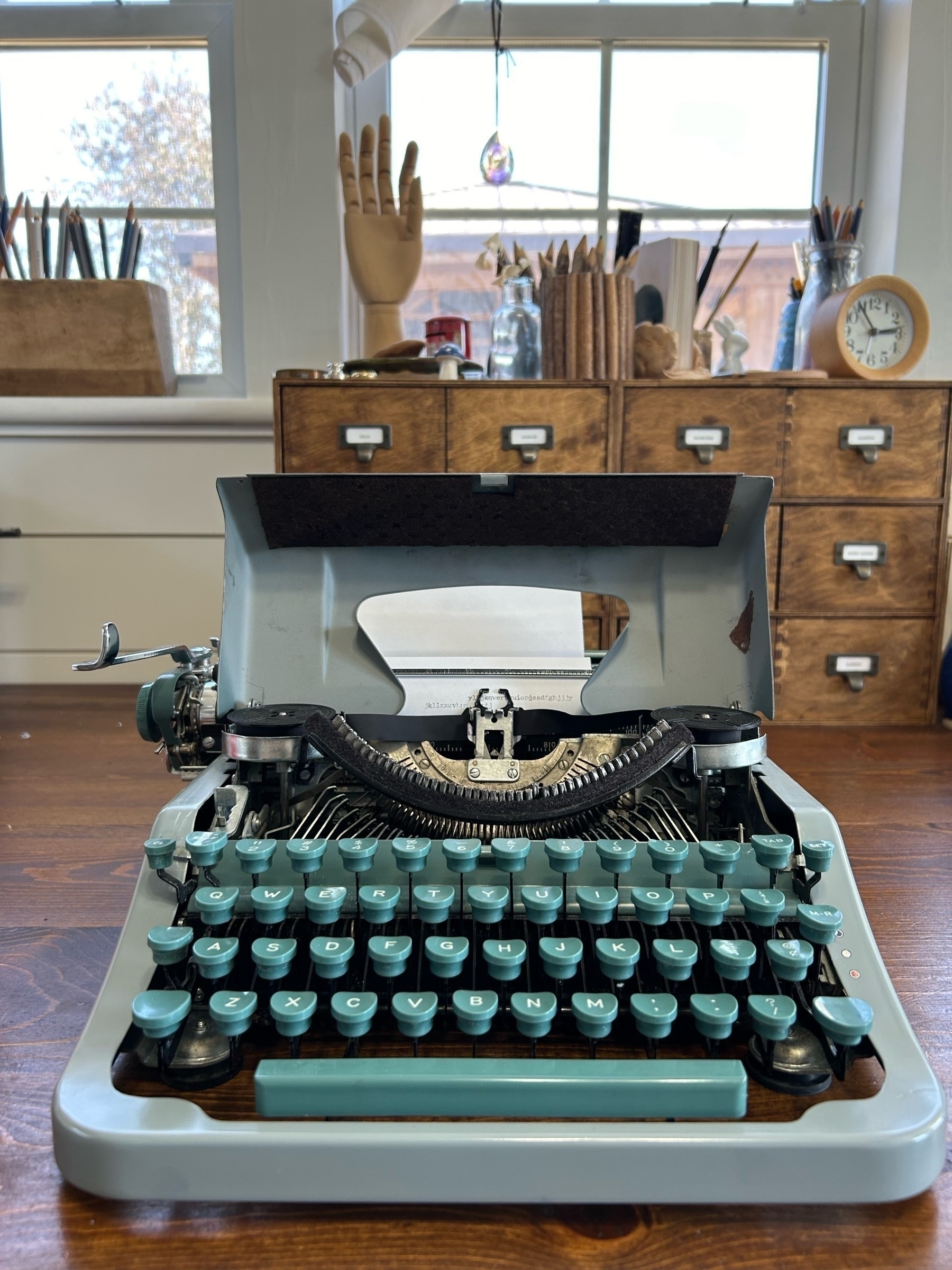 Teal green typewriter with an open carriage for cleaning sits on a wooden desk. Library style drawers and wart supplies are behind.