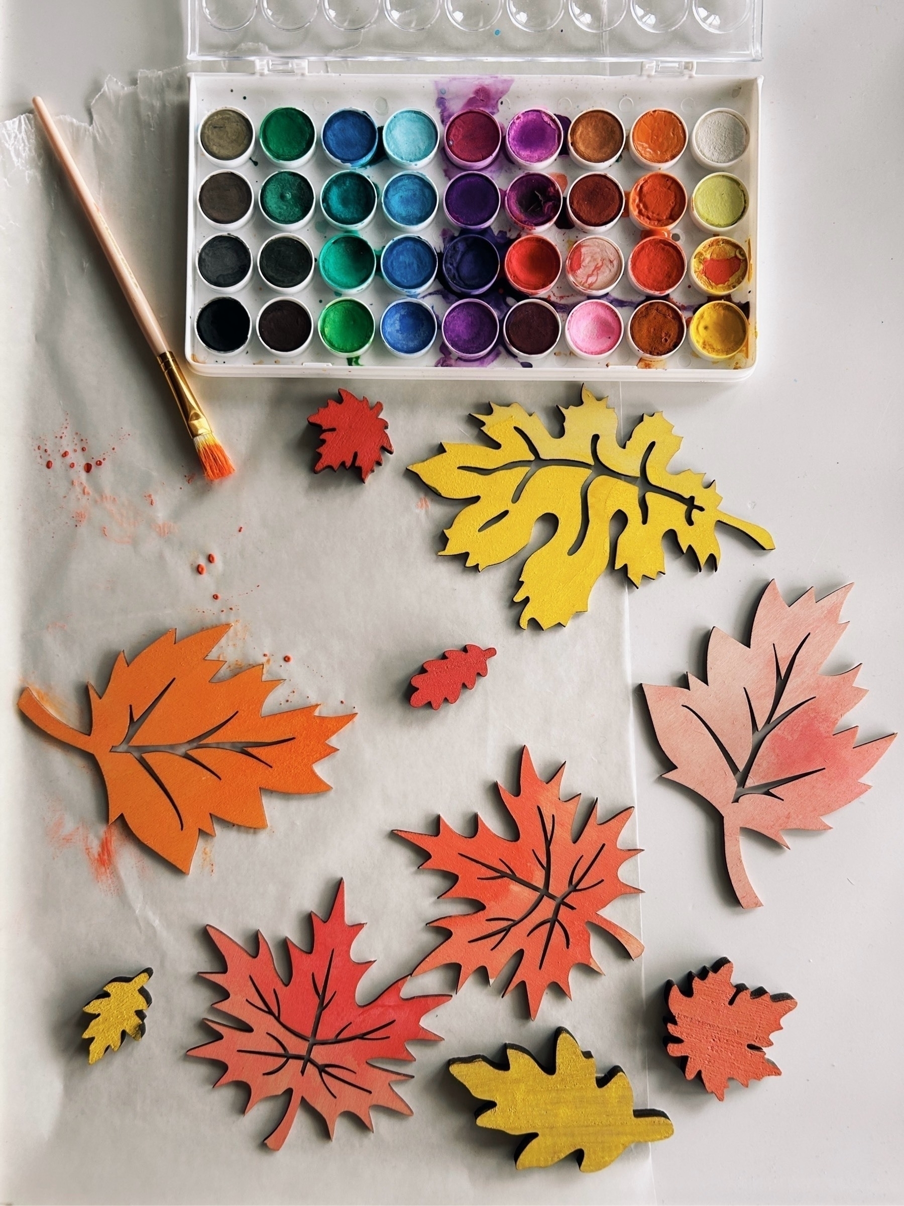 Watercolor paint palette and autumnal painted wooden leaves in oranges and yellow.