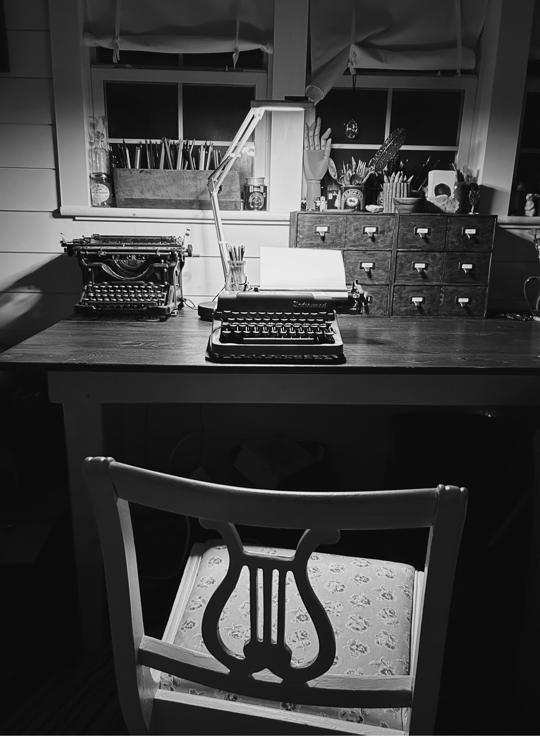 Moody black and white photo of a chair and typewriter at a desk. A second typewriter and library style shelves are beyond.