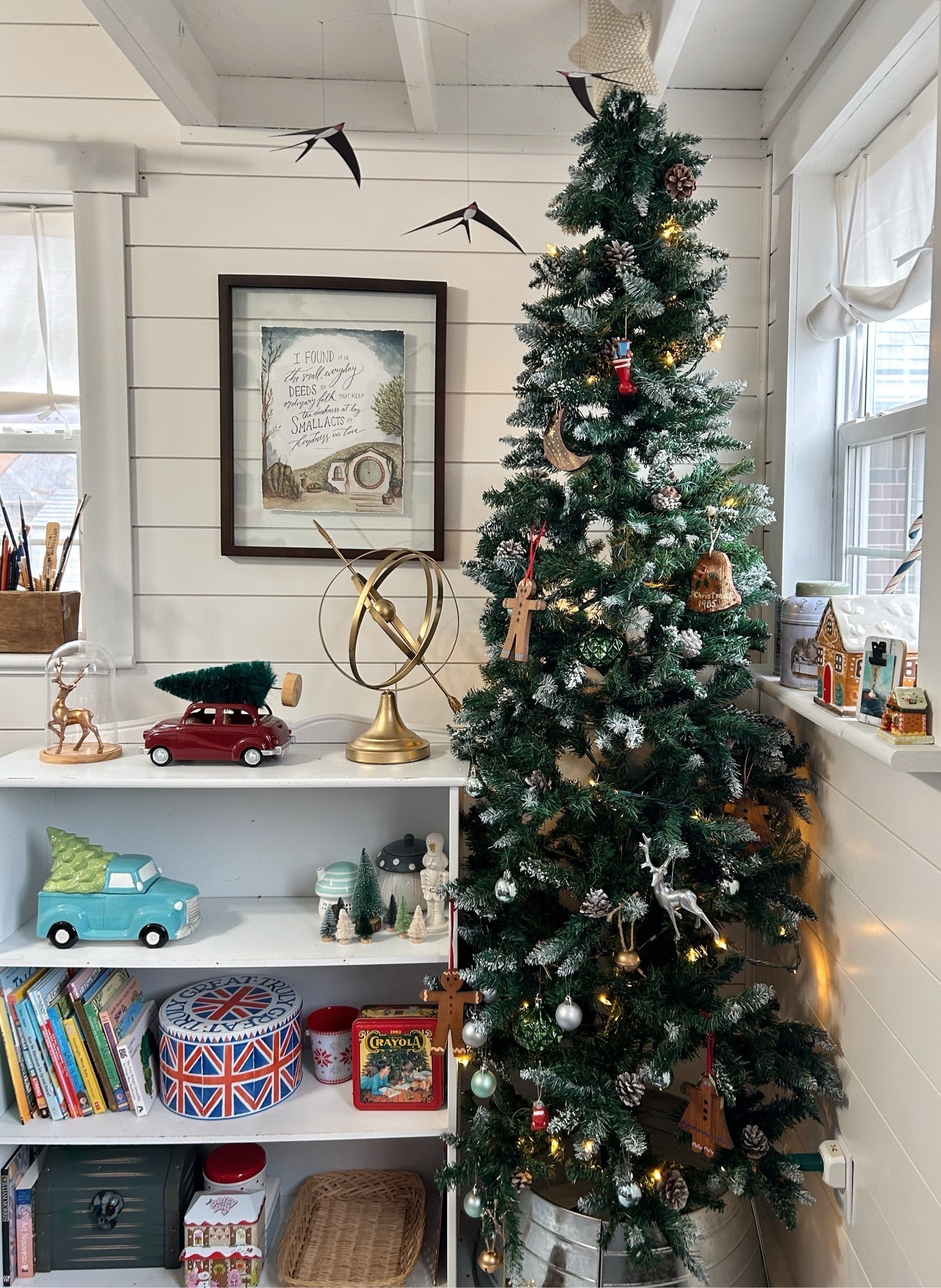 A narrow Christmas tree with wooden ornaments and bells in the corner of a white artist studio. There is a self with trucks, trees, a holden orb, and children's books. On the wall is a hobbit watercolor. In the window are gingerbread houses. A bird mobile hovers above.